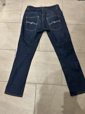 Nudie Jeans Thin Finn Organic Dry 33/32 - Mörkblå jeans från Nudie Jeans, modell Thin Finn Organic Dry. Klassisk femficksdesign med kontrastsömmar och broderade bakfickor. Tillverkade i 98,5% ekologisk bomull och 1,5% elastan för lite stretch. Slimmad passform och raka ben. Jeans läder lappen har tvyärr åkte bort i tvätten men därför säljs dessa för ett billigare pris