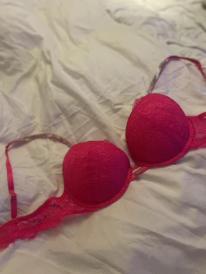Rosa spets-bh med strassdetalj - Säljer en snygg rosa bh med vadderade kupor och spetsdetaljer. Axelbanden är justerbara och har glittrande strass på bägge sidorna. Perfekt för dig som gillar färg och detaljer. Denna Bh är helt ny, och inte använd där den inte passade mig riktigt. 