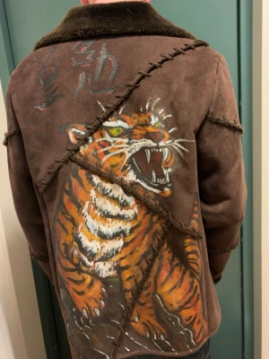 Brun mocka pälsjacka med tigertryck - Unik brun pälsjacka i mocka med grova synliga sömmar och mjuk krage. Baksidan har en stor handmålad tiger och asiatiska tecken, vilket ger jackan en riktigt fet look. Jackan har knäppning framtill och är fodrad med pälsimitation för extra värme. Storlek S,M jag på bilden är 178cm och den passar.