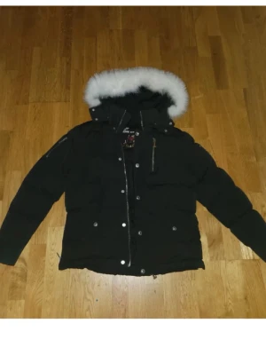 Svart dunjacka från Moose Knuckles - S/m Säljer en svart dunjacka från Moose Knuckles med fluffig vit pälskant på huvan och snygga silverdetaljer. Jackan har dragkedja, knappar och en ikonisk logga på ärmen. Perfekt för kalla dagar och riktigt varm. Flera fickor och quiltad design.
