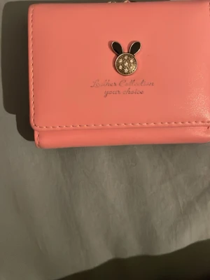 Söt rosa plånbok med kanindekor - Rosa plånbok i syntetmaterial från Luther Collection. Framsidan har en dekorativ kanin-detalj i metall och svart. Plånboken har flera kortfack och ett fönsterfack med blommigt mönster. Perfekt storlek för att få plats i väskan.