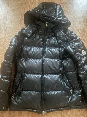 Svart pufferjacka från Moncler - Säljer en svart pufferjacka från Moncler med glansig finish och huva. Jackan har dragkedja framtill, två sidofickor med dragkedja och klassisk Moncler-logga på ärmen. Perfekt för kalla dagar och riktigt snygg till streetwear. Om du har mer frågor de ba säga till! Pris kan diskuteras 