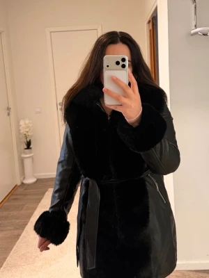 Svart pälsjacka med bälte och dragkedja - Säljer en svart pälsjacka med stora fluffiga detaljer vid krage och ärmslut. Jackan har bälte i midjan och dragkedjor på sidorna. Utsidan är i syntetmaterial och insidan är mjuk och pälsliknande. Perfekt för kalla dagar när du vill vara både varm och snygg. Aldrig använt 