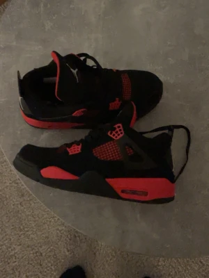 Nike Air Jordan 4 svart/röd sneakers - Säljer ett par Nike Air Jordan 4 sneakers i svart och rött. Skorna har klassisk meshpanel, svarta snören och röda detaljer på sula, mesh och öglor. Ikonisk Jumpman-logga på plösen och hälen. Ovandelen är i syntet och mocka för en clean look.