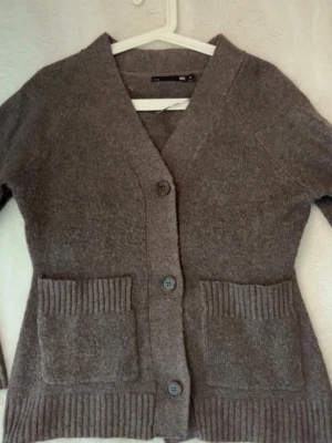 Brun kofta med fickor  - Mysig brun kofta med stora ribbade fickor framtill och tre knappar. Koftan har lång ärm och bred ribbad kant vid ärmslut och nederkant. Perfekt för lager på lager under kyliga dagar. Lite nopprig 