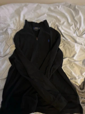 Svart half zip tröja från Polo Ralph Lauren - Klassisk svart långärmad tröja från Polo Ralph Lauren med half zip och blå logga broderad på bröstet. Skön passform och perfekt till lager på lager. Tillverkad i mjuk bomull som känns riktigt nice mot huden.