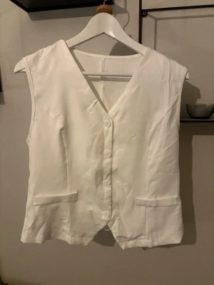 Vit väst med knappar och fickor - Stilren vit väst med v-ringning och fyra knappar framtill. Västen har två dekorativa fickor och är sydd i ett slätt tyg som ger en clean look. Perfekt festtopp eller att ha över skjortan