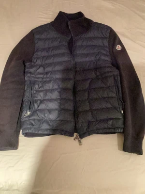 Mörkblå Moncler cardigan med stickade ärmar - Säljer en mörkblå cardigan från Moncler med quiltad front och stickade ärmar. Jackan har hög krage, dragkedja framtill och två fickor med dragkedja. Klassisk Moncler-logga på ärmen och en clean, modern look. Perfekt för dig som vill ha både värme och stil. Den är solblekt men går troligen att lösa genom en kemtvätt osv. Köpt här på Plick.