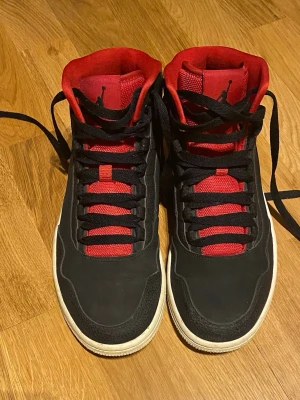 Nike Air Jordan 1 svart/röd sneakers - Säljer ett par Nike Air Jordan 1 sneakers i svart med röda detaljer och snörning. Skorna har en hög modell med rund tå, platt sula och klassisk Jumpman-logga på sidan och tungan. Svarta eller röda snören, ditt val