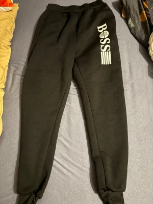 Svarta mjukisbyxor från BOSS - Säljer ett par svarta mjukisbyxor från BOSS med vit logga och grafisk detalj på ena benet. Byxorna har resår i midjan och vid bensluten samt sidofickor. Perfekta för chill dagar eller när du vill ha en clean och enkel look. Pris kan diskuteras!