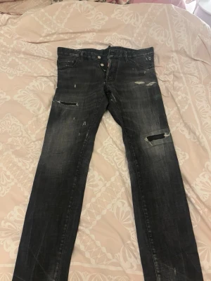 Svarta Dsquared2 Cool Guy Jeans - Säljer ett par svarta Dsquared2 Cool Guy Jeans med slitna detaljer och trasiga partier på benen. Jeansen har knappar i gylfen, fem fickor och en snygg tvättad look. Märke och storlek syns tydligt på etiketten. Perfekta för dig som gillar streetwear.
