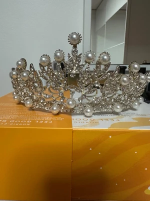Silverfärgad tiara/brudkrona med pärlor och strass - Säljer en riktigt snygg tiara i silverfärg med glittrande strass och stora vita pärlor. Tiaran har ett detaljerat bladmönster och känns lyxig och pampig – perfekt för att ha det på ditt bröllopsdag Sitter stadigt på huvudet och ger en kunglig känsla. Köpt från wedding Castle i Stockholm.