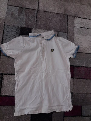 Vit piké från Lyle & Scott Junior - Klassisk vit pikétröja från Lyle & Scott Junior med blå detaljer på krage och ärmslut. Tröjan har korta ärmar, knappslå framtill och den ikoniska gula loggan på bröstet. Tillverkad i mjuk bomull för en skön känsla.