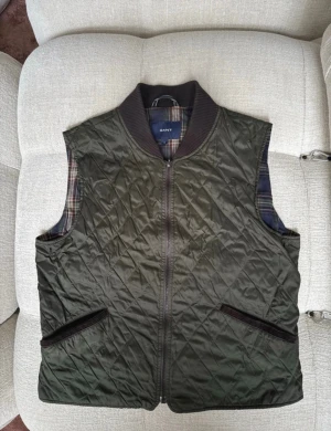 Quiltad grön väst från Gant - Snygg quiltad väst från Gant i mörkgrönt med dragkedja framtill och ribbad krage. Utsidan är i polyester och insidan har ett klassiskt rutigt foder i blått, grönt och rött. Två fickor framtill med mörka detaljer. Perfekt lager på lager-plagg för kyligare dagar.