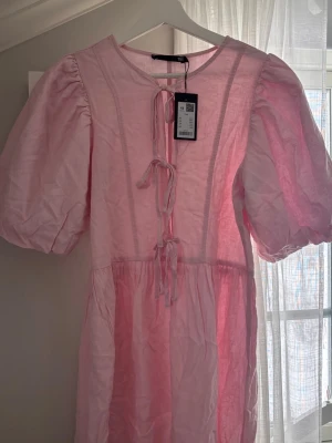 Rosa kortklänning med puffärm och knyt - Supersöt rosa kortklänning från 157 med puffiga korta ärmar och tre dekorativa knyt framtill. Tillverkad i ett lätt och luftigt material som känns perfekt för varma dagar. Klänningen har en avslappnad passform och rund halsringning.