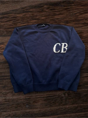 Mörkblå tröja Cole Buxton CB - Mörkblå tröja från Cole Buxton med stora vita CB-bokstäver på bröstet. Klassisk rund halsringning och ribbade muddar vid ärmslut och nederkant. Tillverkad i mjuk bomull för en skön känsla och clean look.