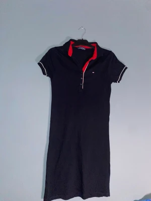 Marinblå pikéklänning Tommy Hilfiger - Snygg marinblå pikéklänning från Tommy Hilfiger med korta ärmar och vit piping vid ärmslut. Klassisk krage med röd insida och knappar framtill. Liten broderad logga på bröstet. Perfekt för en sportig och stilren look.