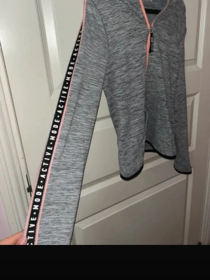 Trårningskofta med luva från H&M barn - Grå tråningskofta med detaljer, fickor, dragkedja och text på ärmarna. Har även klipp till egna Tummhål i ärmarna (se bild) inget som påverkar klädseln, material etc. 