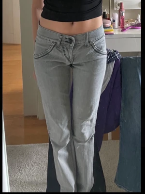 Low waist bootcut jeans - Säljer ett par gråa lågmidjade jeans med bootcut passform. Jeansen har coola detaljer vid fickorna och dubbla knappar i midjan. Materialet är klassisk jeans och färgen är en snygg ljusgrå ton. Perfekta för dig som gillar retrovibbar och vill ha något unikt💝size 25