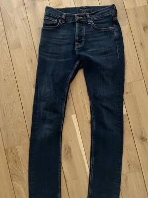 Mörkblå skinny jeans från Nudie Jeans - Säljer ett par mörkblå  jeans från Nudie Jeans i storlek 28/32. Klassisk femficksmodell med orangea sömmar och diskret broderad bakficka. Jeansen har normal passform i midjan och är tillverkade i mjukt jeansmaterial.