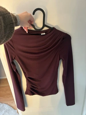 Vinröd långärmad topp med drapering - Säljer en vinröd långärmad topp med snygg drapering över bröstet. Toppen har en tight passform och är tillverkad i ett stretchigt, mjukt material som känns skönt mot huden. Perfekt att styla med jeans eller kjol för en trendig look.