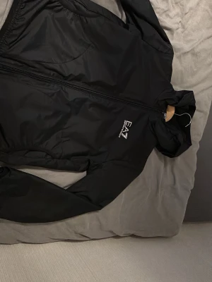 Svart vindjacka från EA7 Emporio Armani - Säljer en svart vindjacka från EA7 Emporio Armani med huva och dragkedja framtill. Jackan har en stilren look med logga på bröstet och är tillverkad i ett lätt och smidigt material. Perfekt för dig som vill ha en snygg och enkel jacka till vardags.