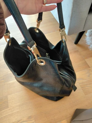 Svart axelväska från Coccinelle - Säljer en svart axelväska från Coccinelle i mjukt skinn med guldfärgade metalldetaljer. Väskan har två rejäla axelband, flera fack och dragkedja. Perfekt för dig som vill ha plats för allt du behöver under dagen.