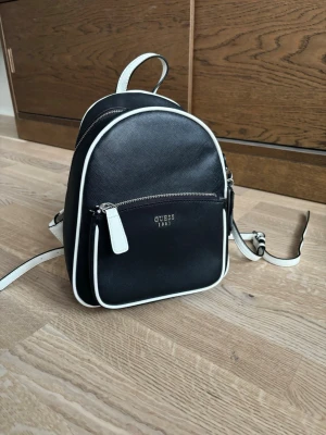 Svart och vit ryggsäck från Guess - Säljer en stilren svart ryggsäck från Guess med vita detaljer och dragkedjor. Väskan har ett litet ytterfack, vita kanter och axelband samt en snygg blomdetalj i svart och vitt. Materialet är syntet med struktur och loggan 'Guess 1981' i silver framtill.