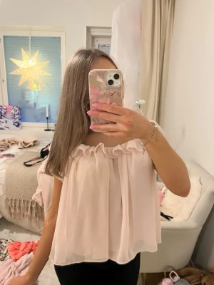 Ljusrosa volangblus i chiffong - Supersöt ljusrosa blus i luftig chiffong med volangdetalj längs den runda urringningen. Blusen är ärmlös och har en flowy, loose passform som ger en romantisk vibe. Perfekt att styla med jeans eller kjol för en söt look.