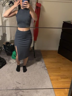 Mörkblå ribbad byxdress med slits - Säljer en mörkblå ribbad byxdress i mjukt stretchigt material. Setet består av ett ärmlöst croppat linne med hög hals och en matchande kjol med slits på sidan. Perfekt för dig som gillar en clean och stilren look.