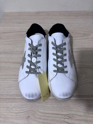 Sneakers från Golden Goose - Snygga vita sneakers från Golden Goose med grå mockastjärna på sidan, grå snörning och svart hälparti. Skorna har platt sula och är tillverkade i skinn med detaljer i mocka. Perfekta för dig som gillar en clean men ändå unik look.