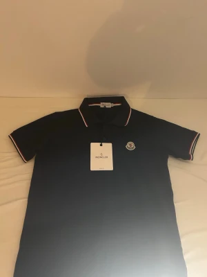 Svart Moncler pikétröja M - Säljer en svart pikétröja från Moncler i storlek M. Tröjan har klassisk krage med röd och vit randig kant, korta ärmar med matchande detaljer och Moncler-logga på bröstet. Tillverkad i bomull för en skön och stilren känsla.