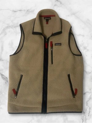 väst från Patagonia - Säljer en beige fleeceväst från Patagonia med svarta detaljer och dragkedjor. Västen har hög krage, tre fickor med dragkedja och röda zip-pullers. Perfekt för lager-på-lager och outdoor-vibe.