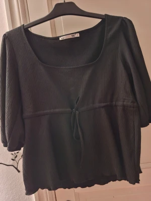 Svart ribbad blus med knyt - Svart blus med ribbad struktur, fyrkantig ringning och trekvartsärmar. Toppen har en söt knytning framtill i midjan och lätt puffade ärmar. Materialet är mjukt och stretchigt, perfekt för en chill och stilren look.