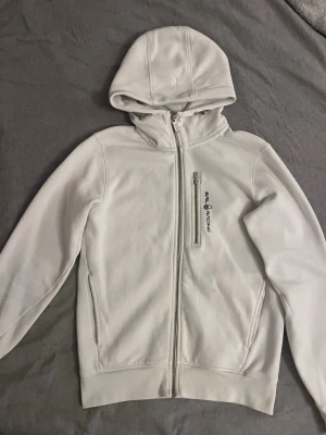 Sail racing zip hoodie  - En tröja med en dragkedja framtill och en huva. Den har en ficka på bröstet och två fickor framtill. Varumärket "Sail Racing" är tryckt på vänster bröst. Den är väldigt varm och stilren.