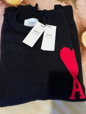 Svart stickad tröja från Ami - Säljer en svart stickad tröja från Ami Alexandre Mattiussi med rund hals och ribbade muddar. Tröjan har en stor röd hjärtapplikation med bokstaven A på bröstet, vilket ger en unik och trendig look. Perfekt för dig som gillar stilrena plagg med en twist. M/L passar