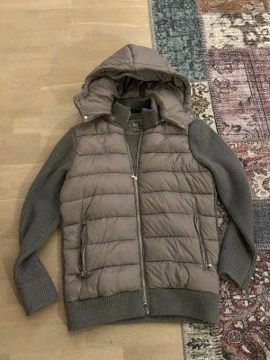 Grå pufferjacka från Leon Marcel - Säljer en grå pufferjacka från Leon Marcel i storlek M. Jackan har stickade ärmar och rygg, quiltad front och avtagbar huva. Två fickor med dragkedja framtill och ribbade muddar för extra komfort. Perfekt för kyliga dagar.