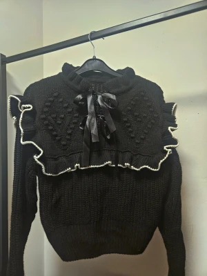 Svart stickad tröja med volang och rosett - Supersöt svart stickad tröja med volangdetaljer och vit kant längs volangen. Framtill finns en stor rosett i satinband och ett upphöjt mönster på bröstet. Tröjan har lång ärm och ribbad hals. Använd typ 1 gång så väldigt fint skick.