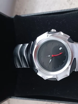 Unik klocka med "Alien" Design - Säljer en riktigt häftig klocka med en design som sticker ut.                                                           Design: Silverfärgad länk med "ryggradskänsla" och en svart urtavla med ett unikt, vågigt mönster i mitten. Detaljer: Den klarröda sekundvisaren ger en snygg kontrast mot det svarta och silvriga. Har även datumvisning. Skick: I använd skick. Stil: Perfekt för dig som gillar cybercore, gorpcore eller unika Y2K-accessoarer.