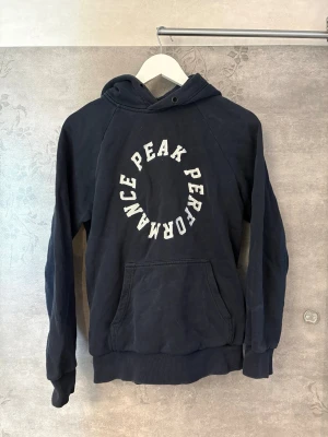 Mörkblå Peak Performance hoodie - Snygg mörkblå hoodie från Peak Performance med vit text i cirkelformad design på bröstet. Klassisk känguruficka framtill, ribbade muddar och huva. Perfekt för en avslappnad och sportig stil. Materialet känns mjukt och bekvämt.
