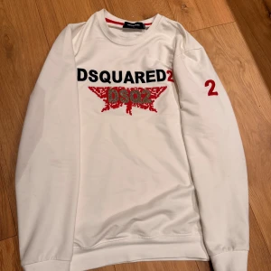 Vit sweatshirt från Dsquared2 - Snygg vit sweatshirt från Dsquared2 med svart och rött tryck framtill samt röd siffra på ärmen. Rund halsringning och ribbade muddar. Perfekt för dig som gillar streetwear och vill sticka ut med ett exklusivt plagg.