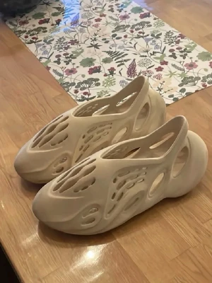 Beige adidas Yeezy Foam Runner tofflor - Säljer ett par beige adidas Yeezy Foam Runner tofflor med futuristisk design och många utskurna detaljer. Tillverkade i syntetmaterial som ger en lätt och bekväm känsla. Perfekta för dig som vill ha något unikt och trendigt på fötterna.
