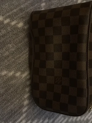 Lv väska - Snygg handväska från Louis Vuitton i klassiskt brunt rutigt mönster. Väskan har en kompakt form och är tillverkad i skinn med mörka och ljusare bruna rutor. Perfekt för dig som gillar lyxiga accessoarer med ikonisk design.