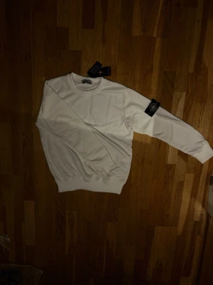 Vit sweatshirt från Stone Island - Snygg vit sweatshirt från Stone Island med klassisk rund hals och ribbade muddar. Tröjan har den ikoniska Stone Island-loggan på vänster ärm och är gjord i mjukt bomullsmaterial. Perfekt för en clean och stilren look. Kan passa en storlek mindre och kanske en större också. Skriv om ni undrar något!