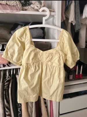 Ljusgul puffärm topp i bomull - Söt ljusgul topp med puffärmar och fyrkantig ringning. Tillverkad i mjuk bomull och har en avslappnad passform med rynkade detaljer under bysten. Perfekt för dig som gillar en feminin och trendig stil.