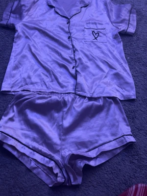 Lila satinpyjamas med korta ärmar - Snyggt pyjamasset i glansigt lila satinmaterial med kortärmad skjorta och matchande shorts. Skjortan har svarta knappar, krage och en bröstficka med ett broderat hjärta. Shortsen har elastisk midja och svarta kantdetaljer. Perfekt för mysiga kvällar hemma.