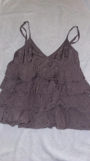 Lila volanglinne med tunna band H&M - Säljer ett lila linne med tunna axelband och flera volanglager. Tyget är lätt och har en skrynklig look som ger en avslappnad vibe. Perfekt för sommardagar eller att styla lager på lager.