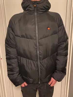 Svart pufferjacka från Ellesse - Snygg svart pufferjacka från Ellesse med huva och dragkedja framtill. Jackan har en quiltad design och en liten logga på bröstet. Perfekt för kalla dagar och ger en riktigt chill streetwear-look.