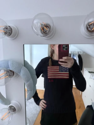Mörkblå stickad tröja Ralph Lauren - Mörkblå stickad tröja från Ralph Lauren med amerikansk flagga och RL-broderi på bröstet. Tröjan har rund halsringning, långa ärmar och ribbstickade muddar vid ärmslut och nederkant. Perfekt för en klassisk och stilren look.