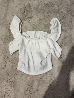 Vit offshoulder topp med volang - Superfin vit offshoulder topp med bred volang runt axlarna. Toppen är croppad och har en lätt ribbad struktur i tyget. Perfekt för dig som gillar en söt och trendig stil.